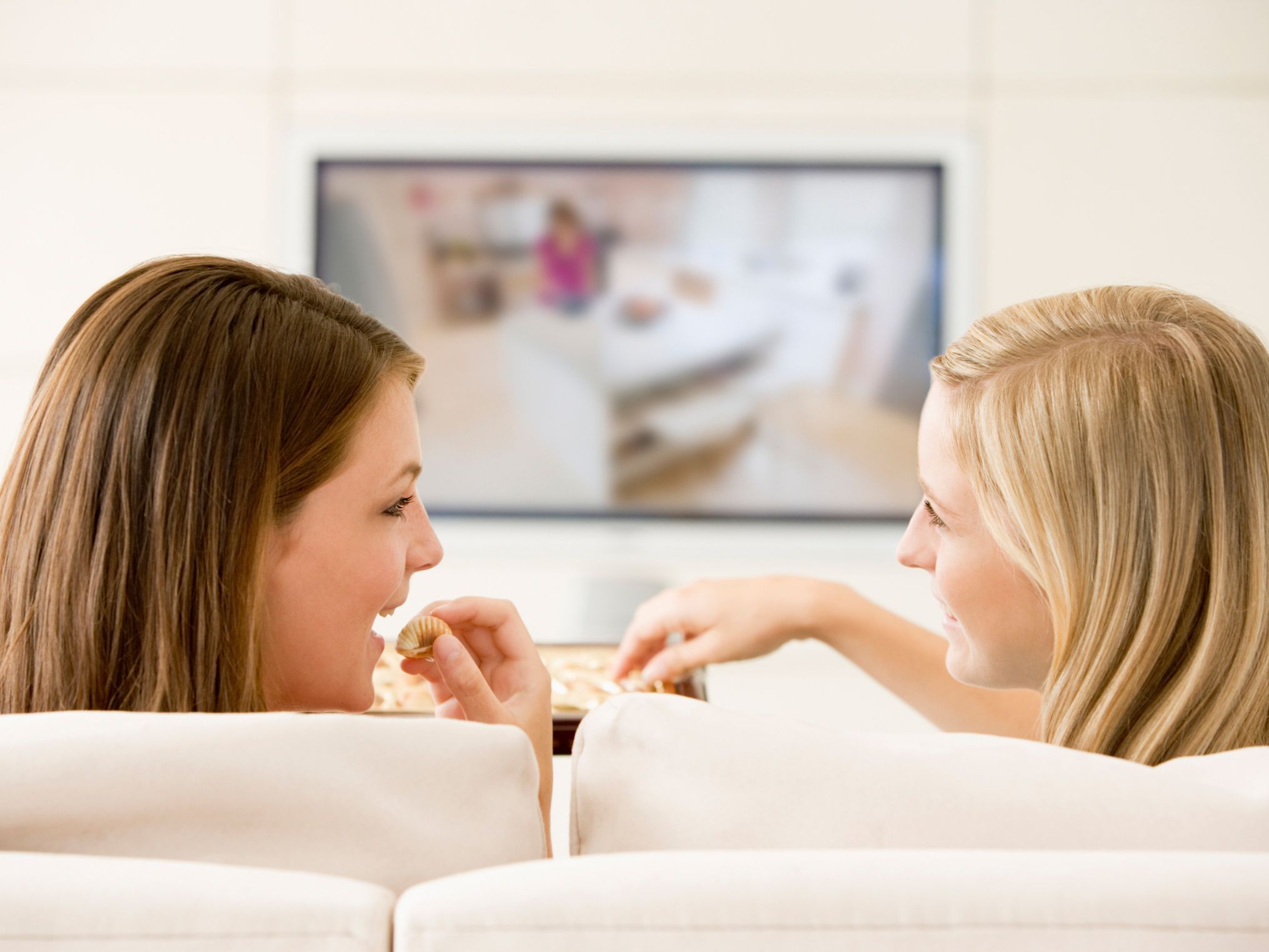 Two women in living room watching television eating chocolates s